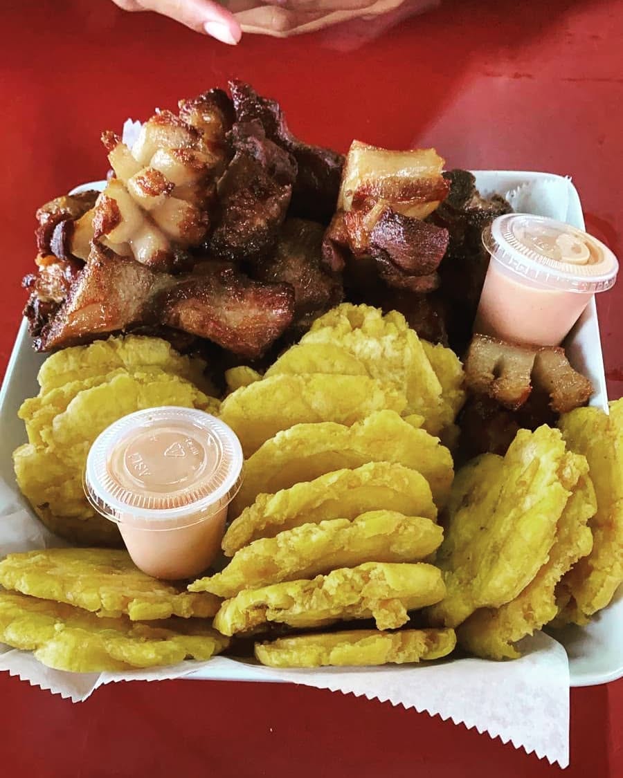 Bandeja con carne frita crujiente, tostones dorados y dos salsas rosadas sobre mesa roja